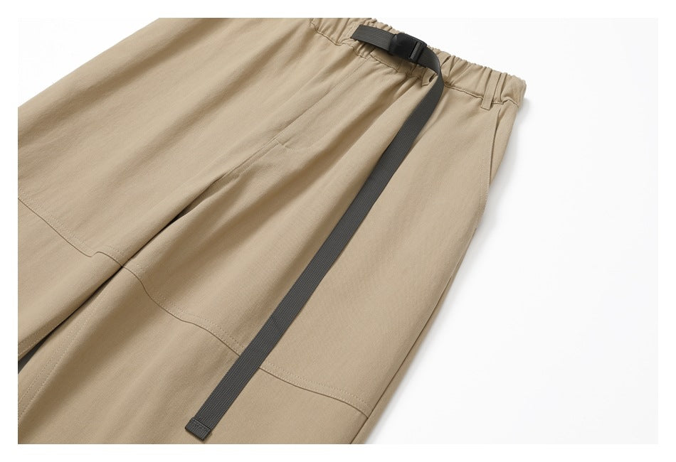 Partial flat lay of tan pants with an elastic waist, dark webbing belt, and stitched panel details.