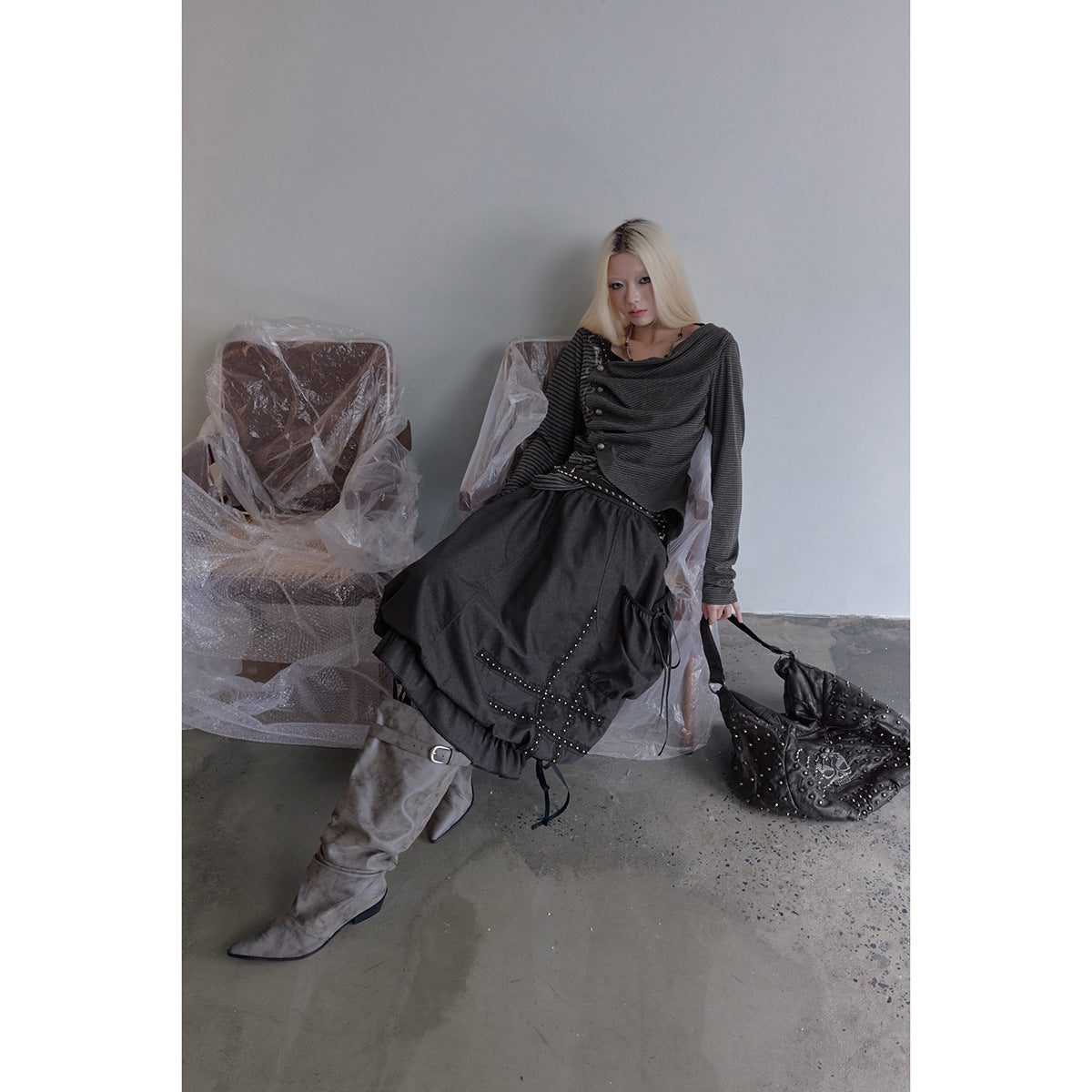 Model seated in a charcoal gathered skirt with studded trim and ties, a striped top, and gray boots.