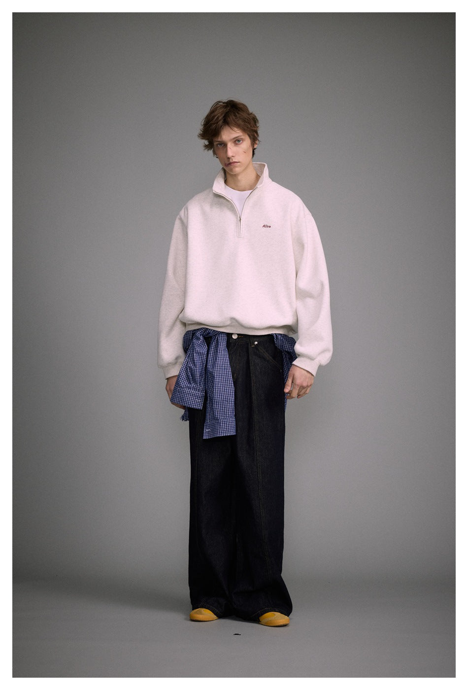 Model wearing a light heather quarter-zip sweatshirt with a raised collar, styled with wide black jeans and a blue checked