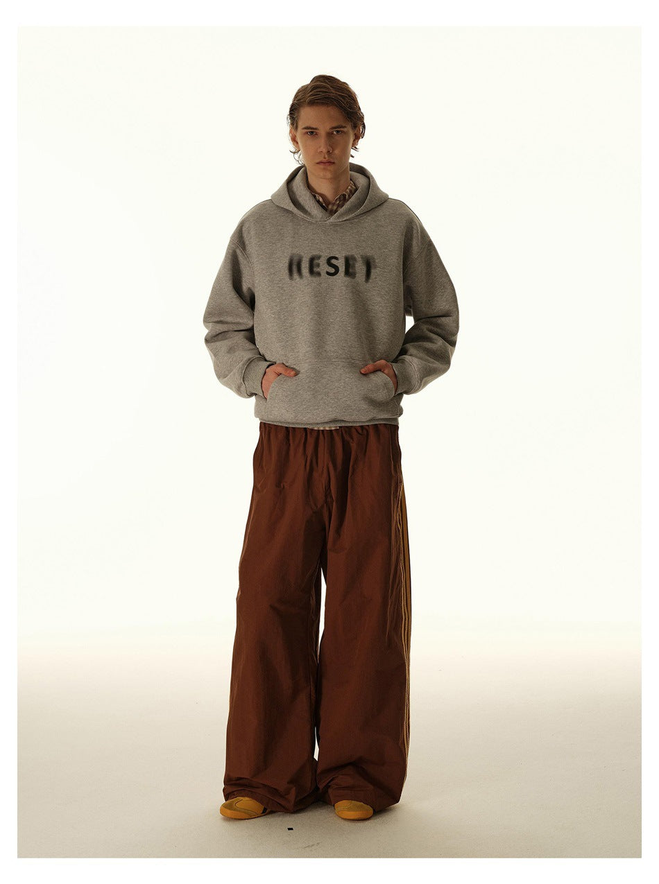 Model in a gray printed hoodie and loose brown track pants with narrow side stripes.