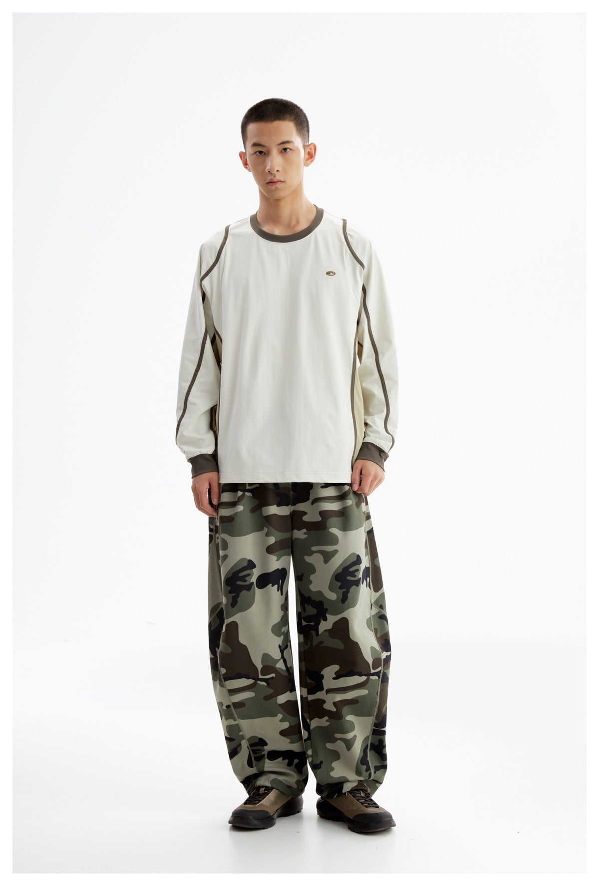 Front view of a model wearing an off-white long-sleeve shirt with dark contrast piping and loose green camouflage pants.