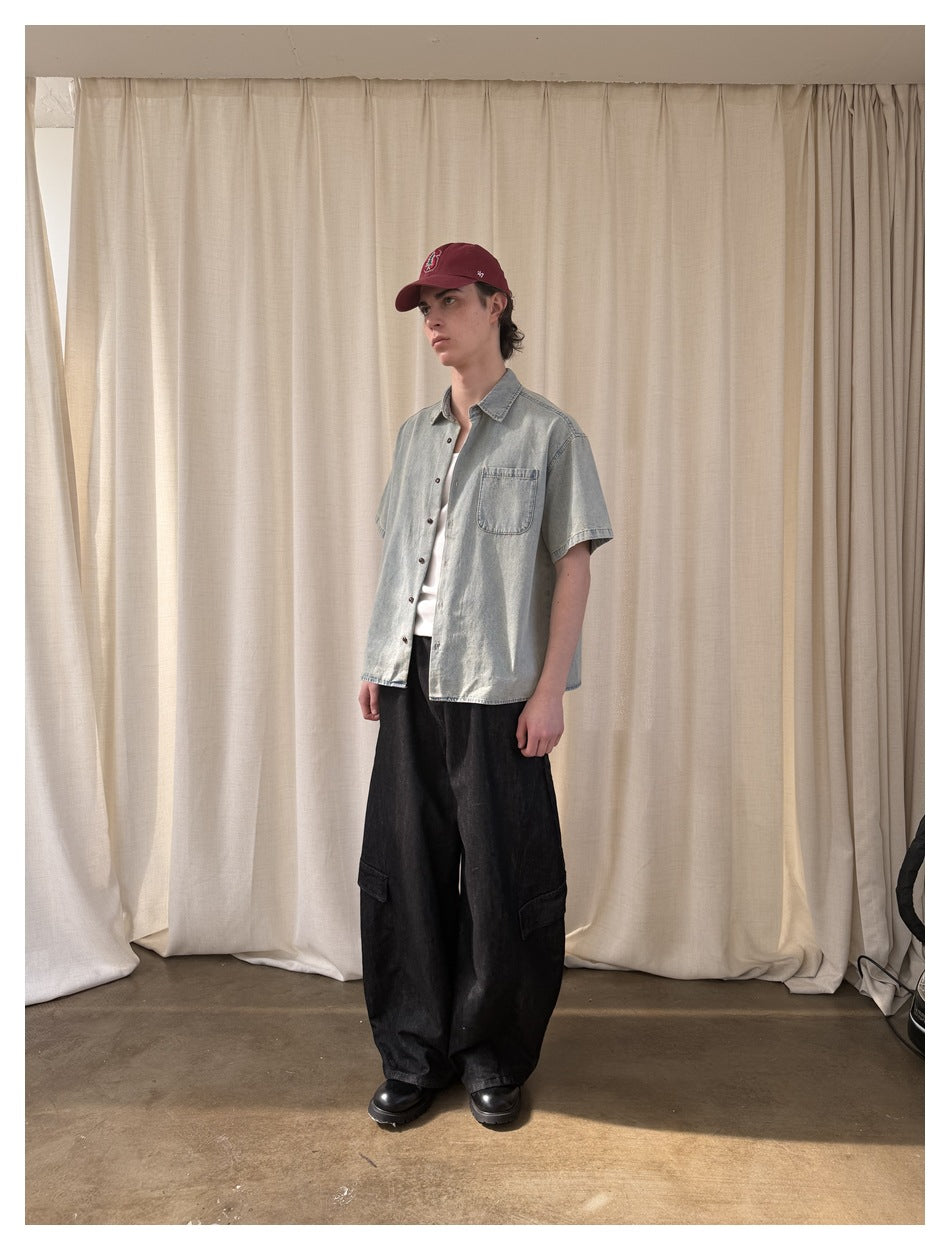 Model wearing washed light blue short-sleeve shirt open over white tee with black wide cargo pants and red cap