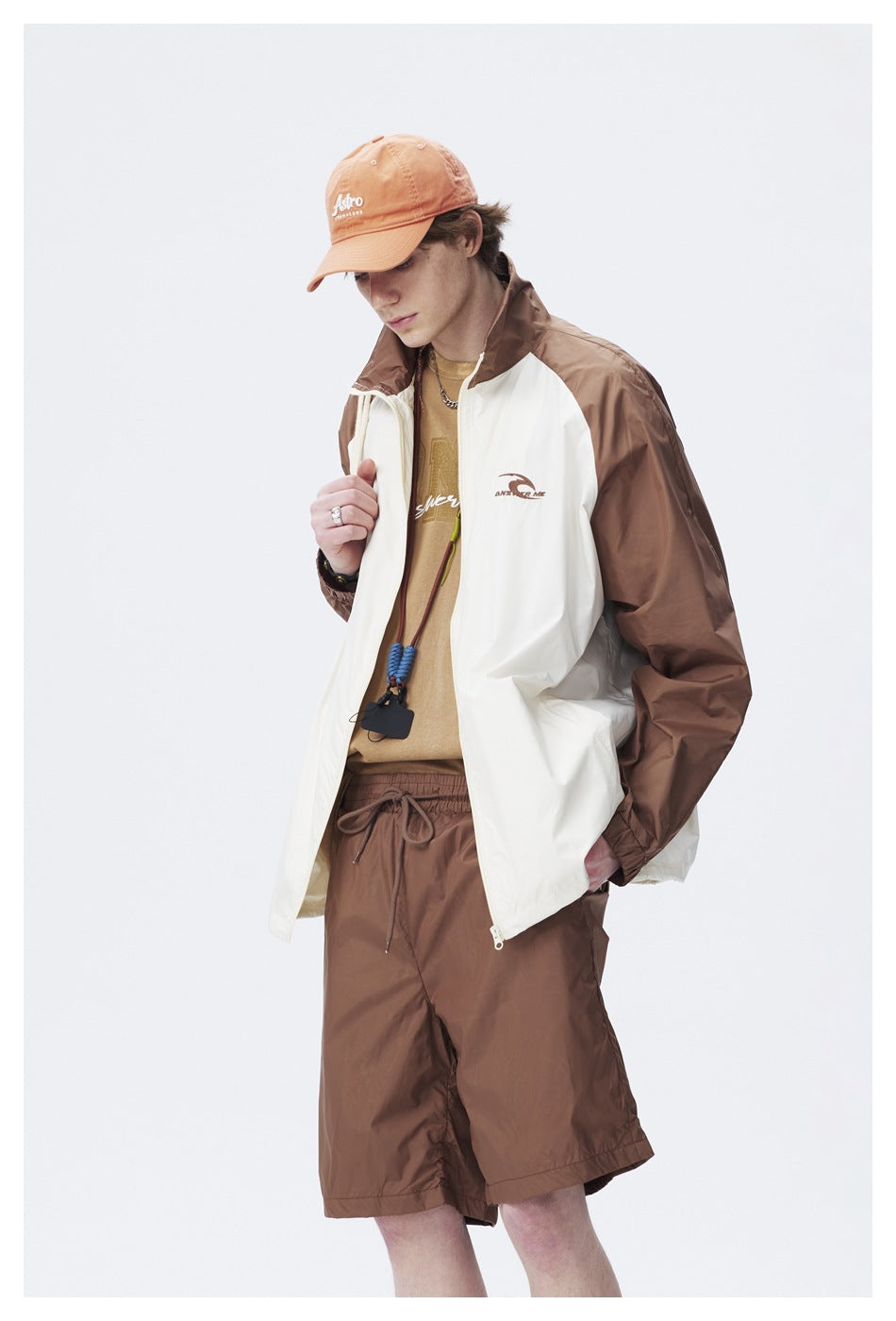 Model wearing a cream-and-brown color-block zip jacket and brown shorts, with an orange cap.