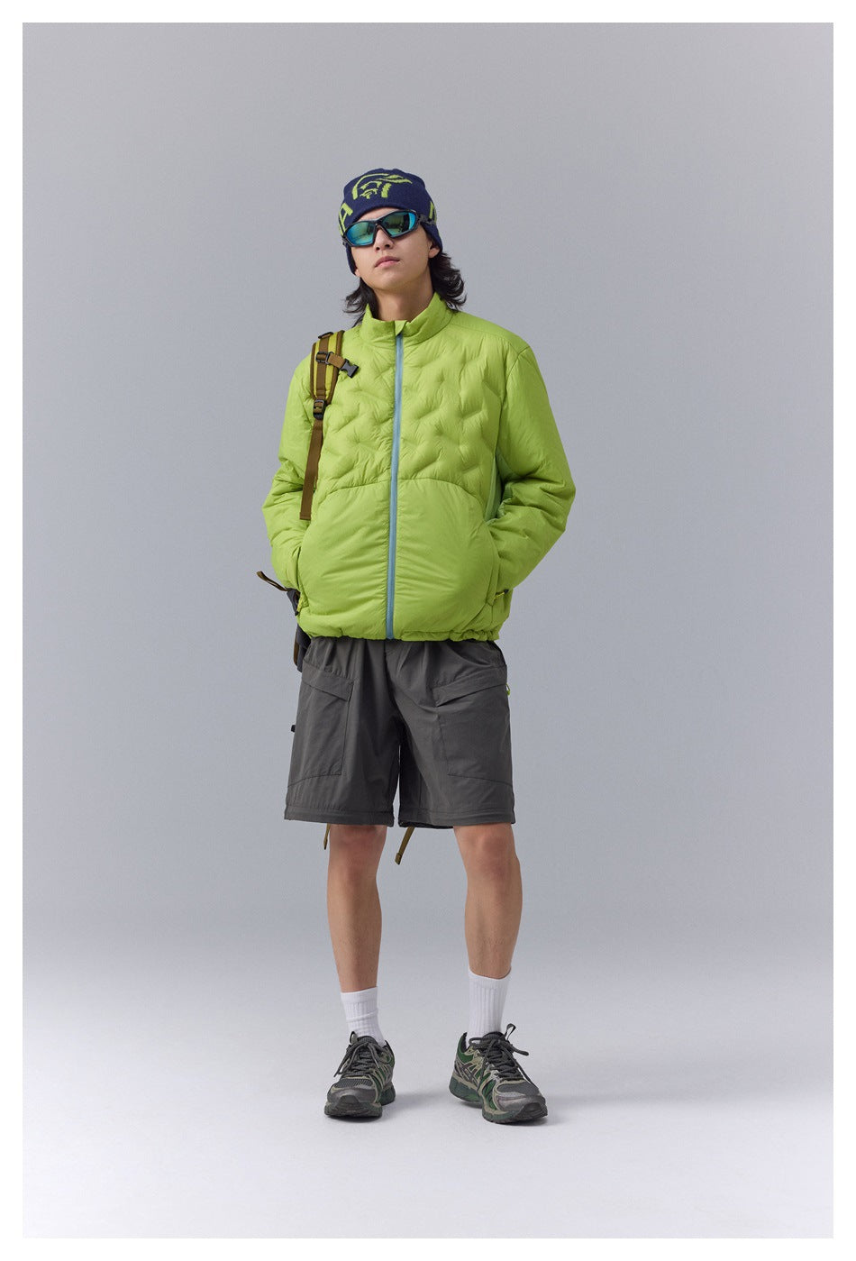 Model standing in a lime green quilted zip jacket with gray cargo shorts, beanie, sunglasses and a backpack strap