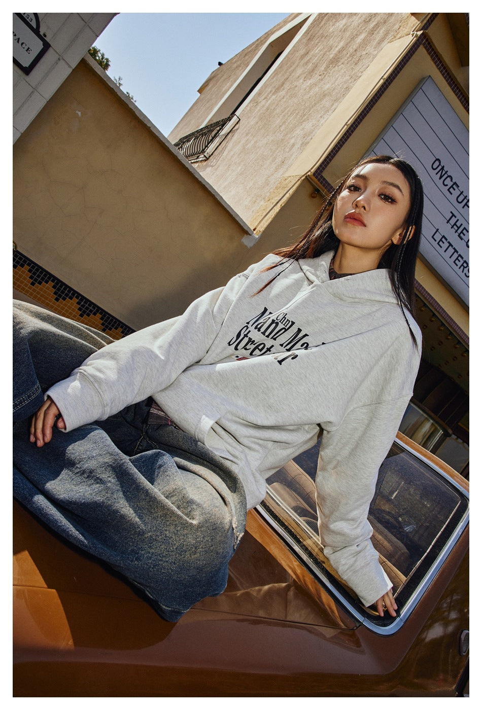 A woman sits on a car wearing a light gray hoodie with black “Hand Made Street wear” lettering and blue jeans.