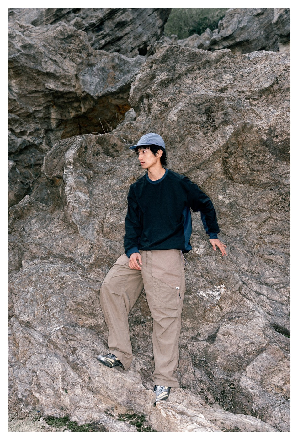 Model seated on rocks wearing a dark navy long-sleeve top, taupe cargo pants, and a gray cap.