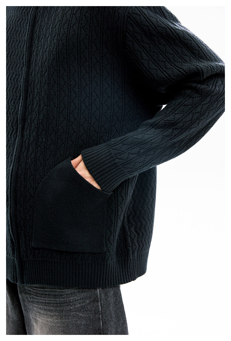 Cropped side view of a model wearing a black textured knit sweater, showing its sleeve cuff and large front pocket.
