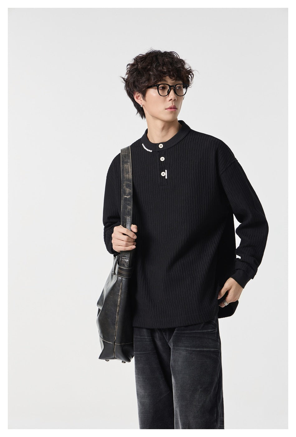 Person wearing a black ribbed Henley top with a three-button placket, holding a leather shoulder bag, upper-body view