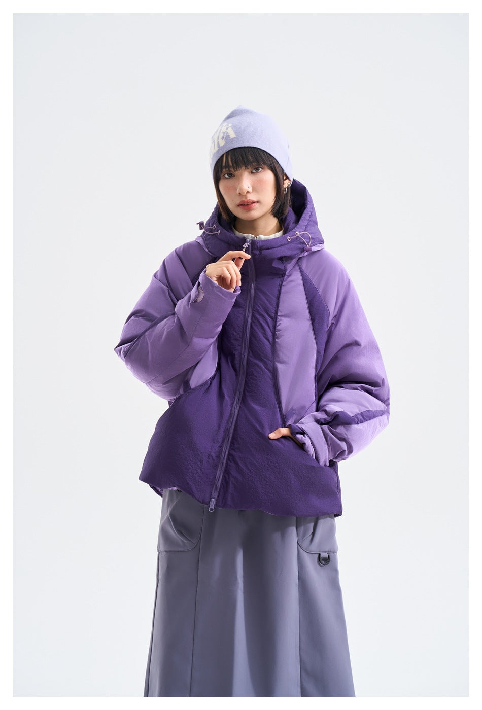 Model wearing a purple two-tone hooded puffer jacket, hand on zip, with lavender beanie and gray skirt