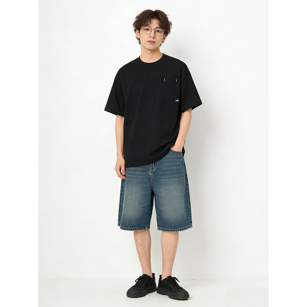 Model wearing a black short-sleeve T-shirt with a chest pocket and blue denim shorts.