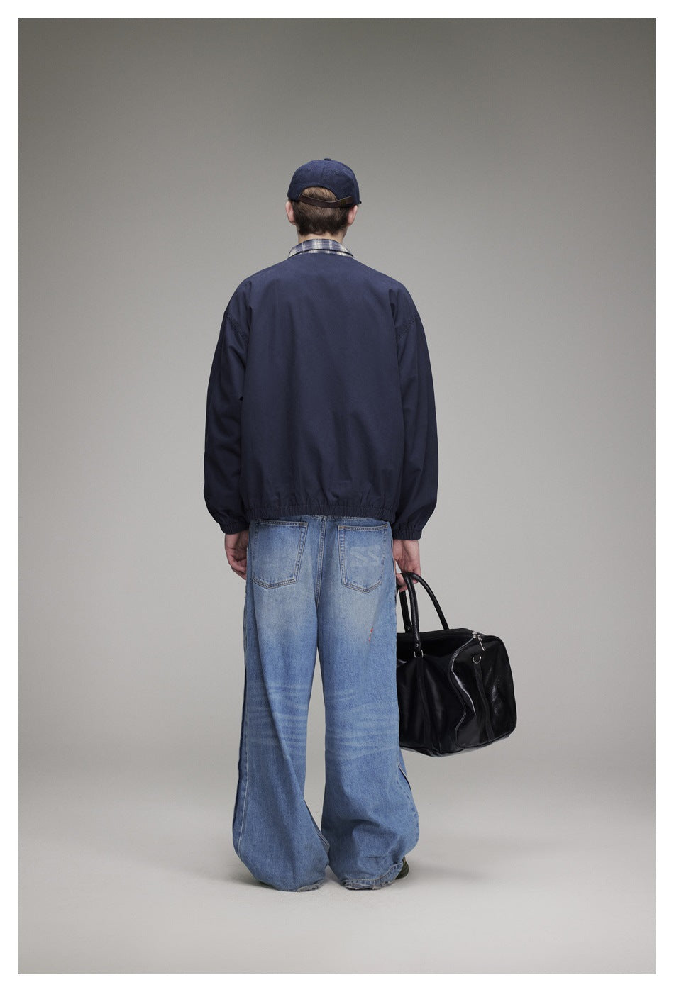 Back view of model in navy jacket with elastic hem, wide-leg jeans, cap and black duffel bag