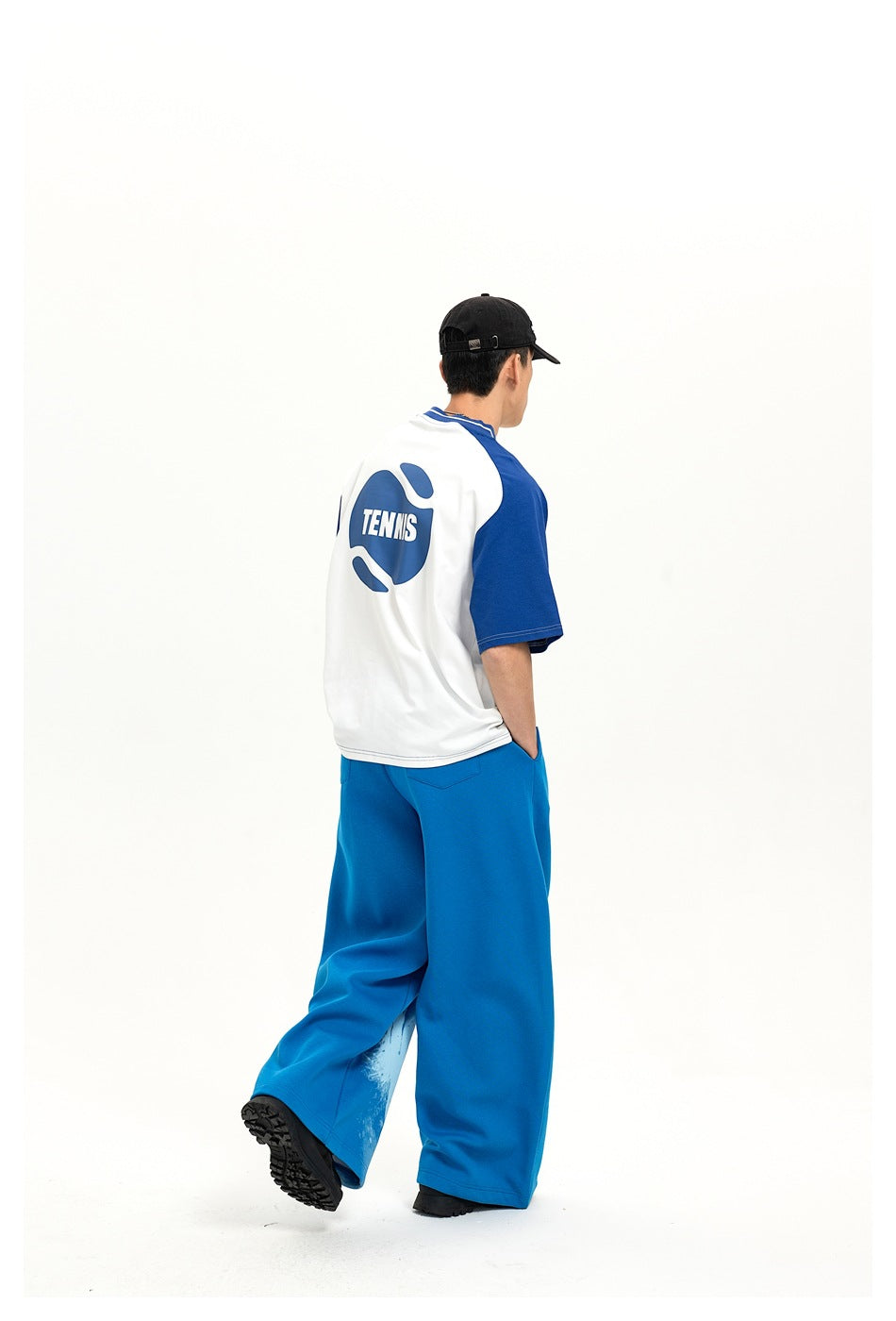Rear three-quarter view of a model wearing blue pants and a white-and-blue shirt with a large tennis graphic.
