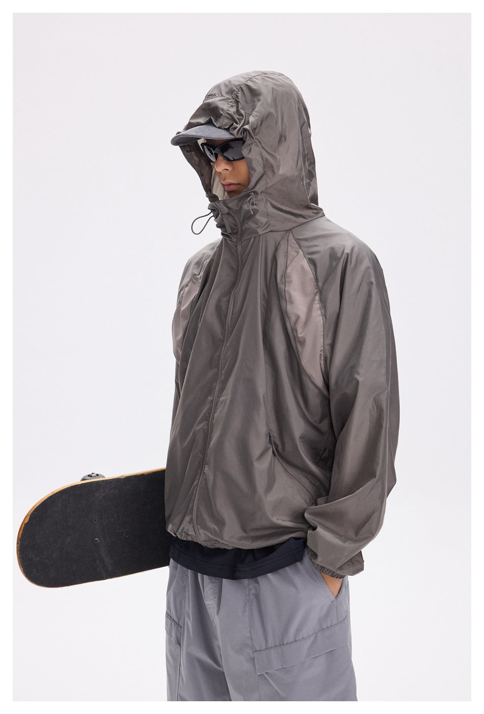 Model wearing a dark taupe-gray hooded zip jacket with sunglasses and carrying a skateboard.