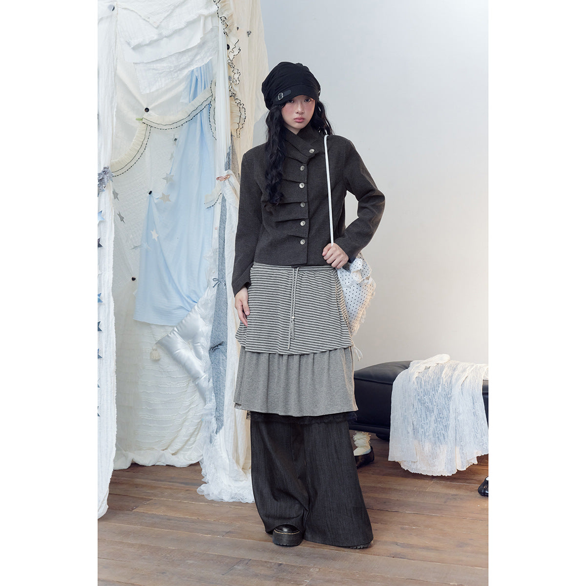 Front view of a model in a charcoal buttoned jacket, gray striped layered skirt, and wide dark pants.