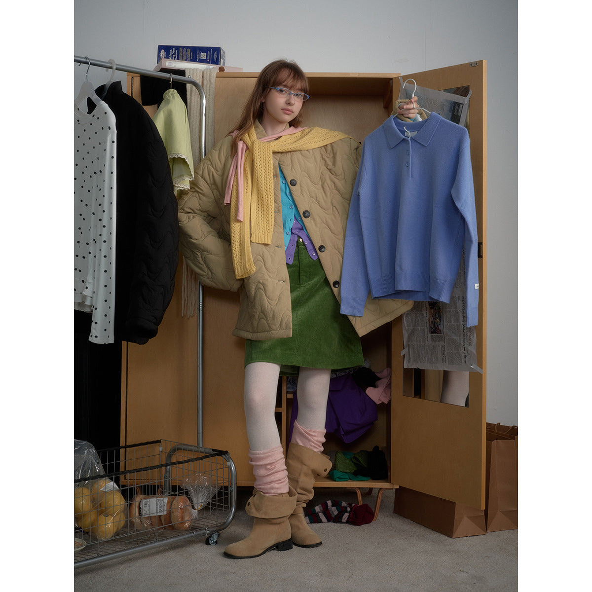 Model wearing a beige wave-quilted coat, mustard scarf and green skirt while holding a blue collared sweater.