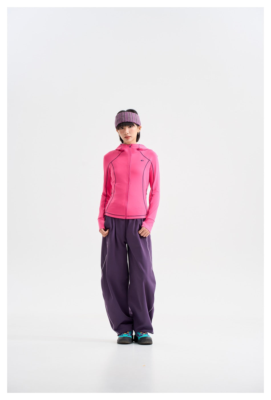 Model wearing a bright pink zip-front hooded jacket with dark piping, loose purple pants, and a purple headband.