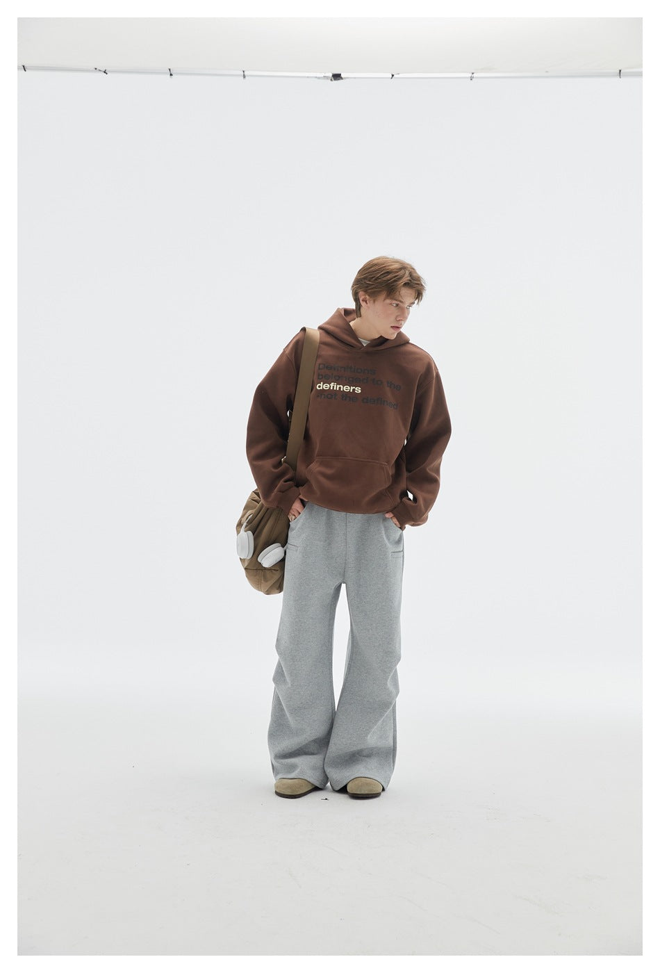 Full-length model in a brown text-print hoodie and loose gray pants, carrying a shoulder bag.