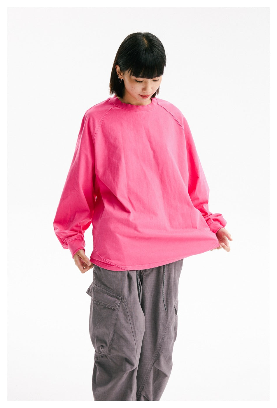 Model looks down and holds the hem of a bright pink oversized crewneck, paired with gray cargo pants.