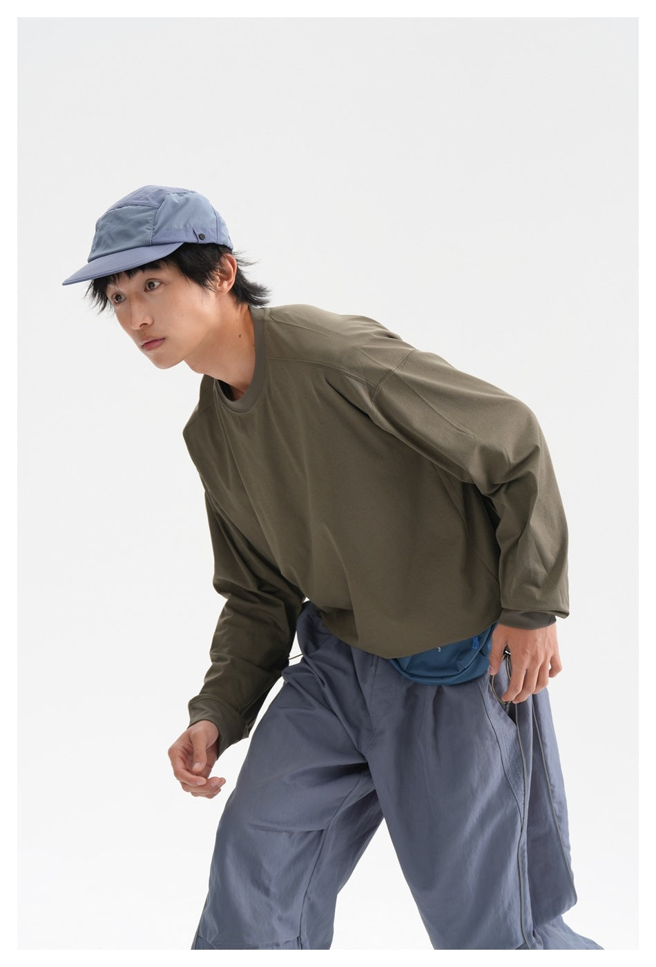 Man leaning forward in an olive long-sleeve shirt and slate-blue trousers, wearing a blue cap and waist bag.