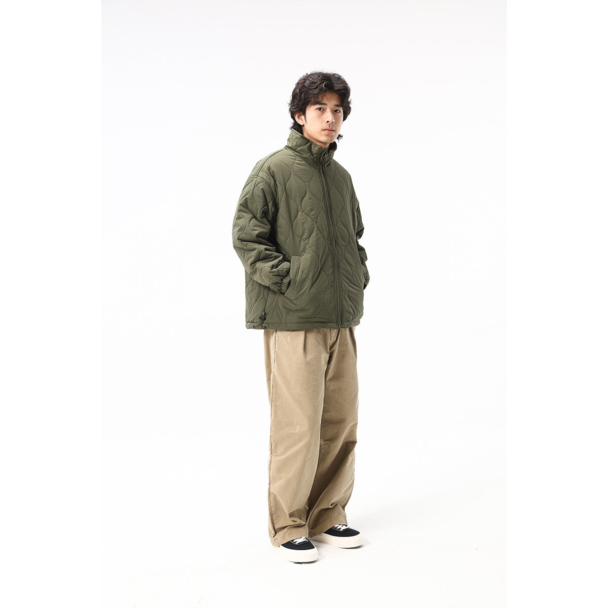 Full-length view of a man wearing an olive quilted zip jacket, loose tan pants and black-and-white sneakers.