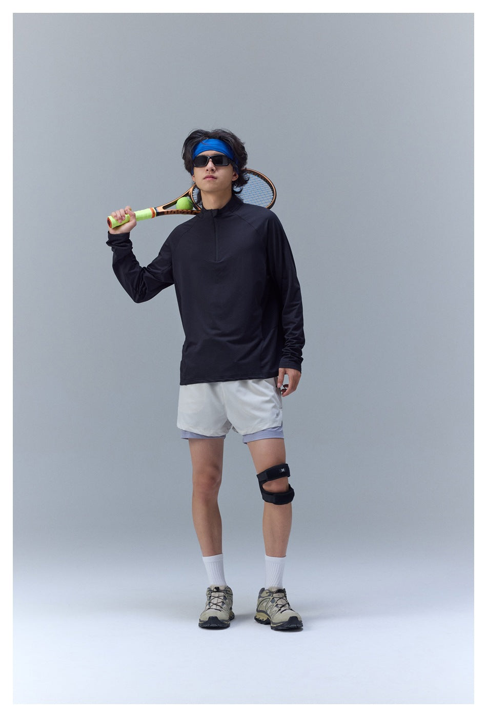 Model in a black long-sleeve quarter-zip tennis top and light shorts, holding a racket over his shoulder.