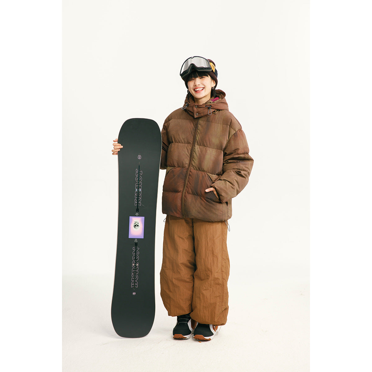 Model wearing a brown hooded puffer jacket and loose brown pants beside a black snowboard.