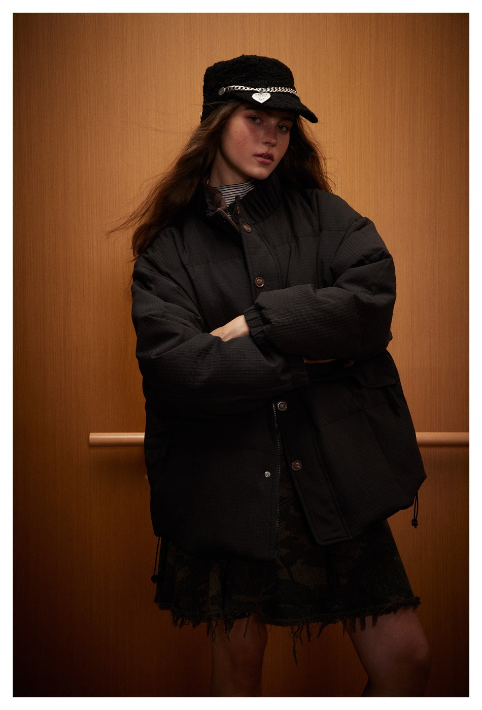 Model wearing a black quilted jacket with a high collar and snap-front closure, paired with a dark patterned skirt.