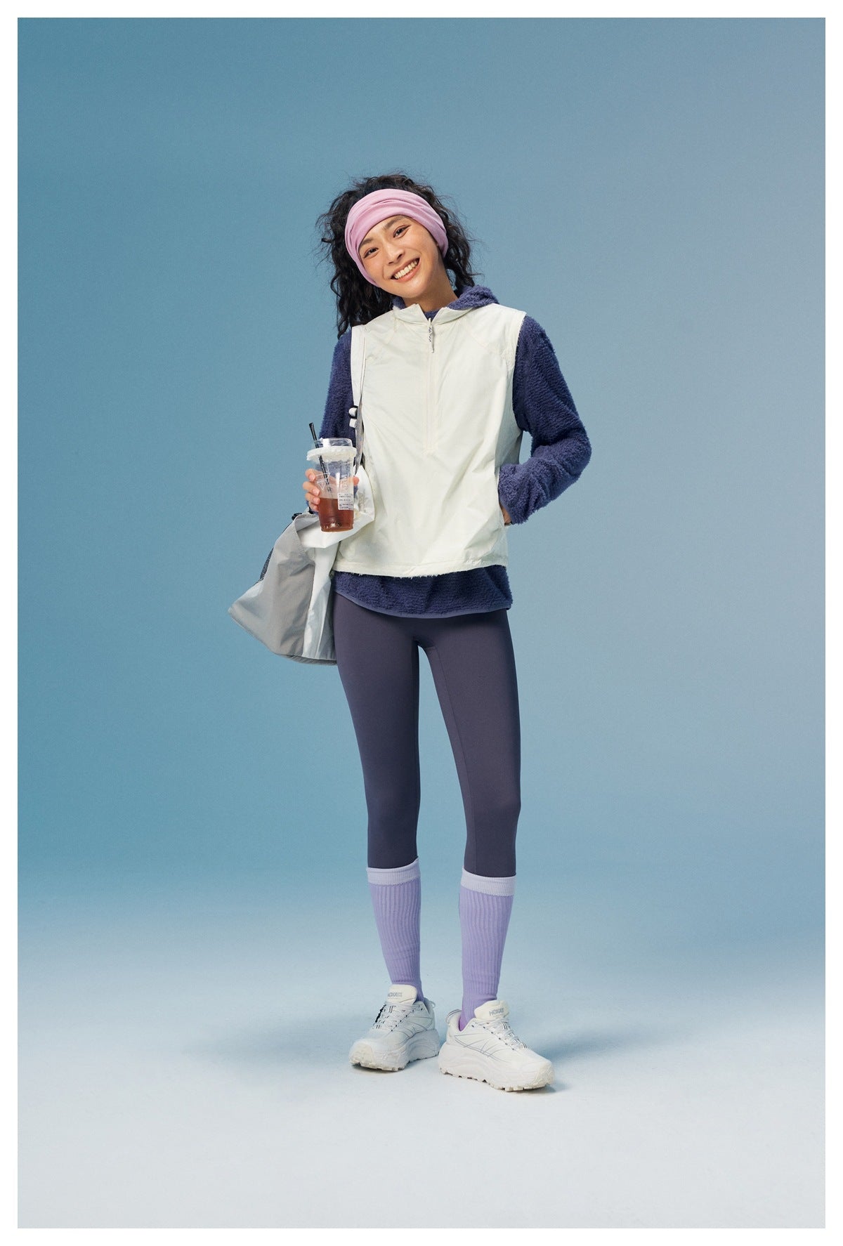 Woman modeling a white zip-front vest over a navy fuzzy hooded top, with purple leggings and a pink headband.