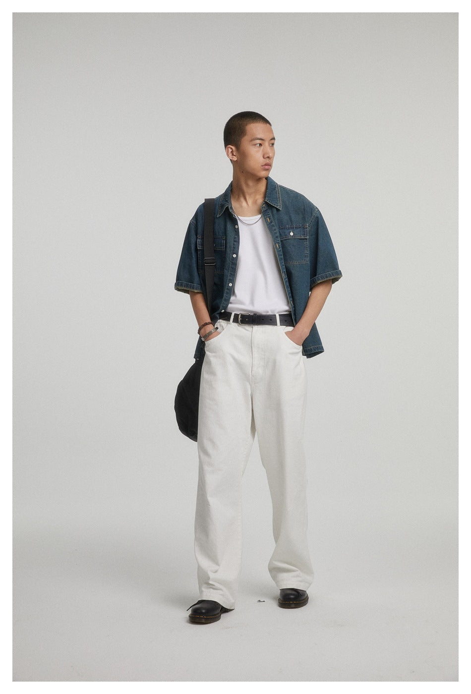 Model wearing a short-sleeve dark blue denim button-up shirt over a white tank, with white trousers and a black shoulder bag.