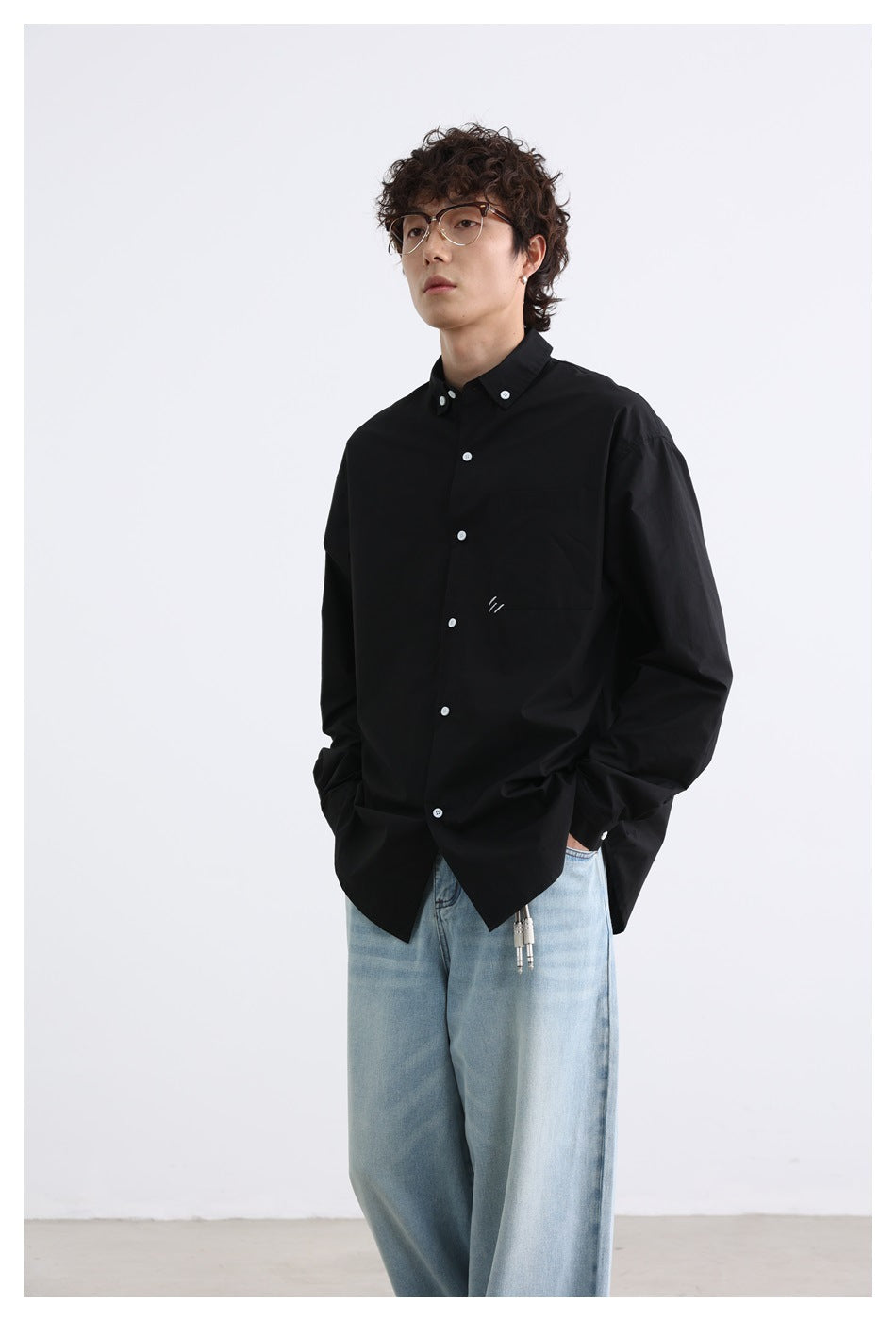 Model wearing a loose black button-down shirt with a chest pocket, white buttons, and a small white stitched mark.
