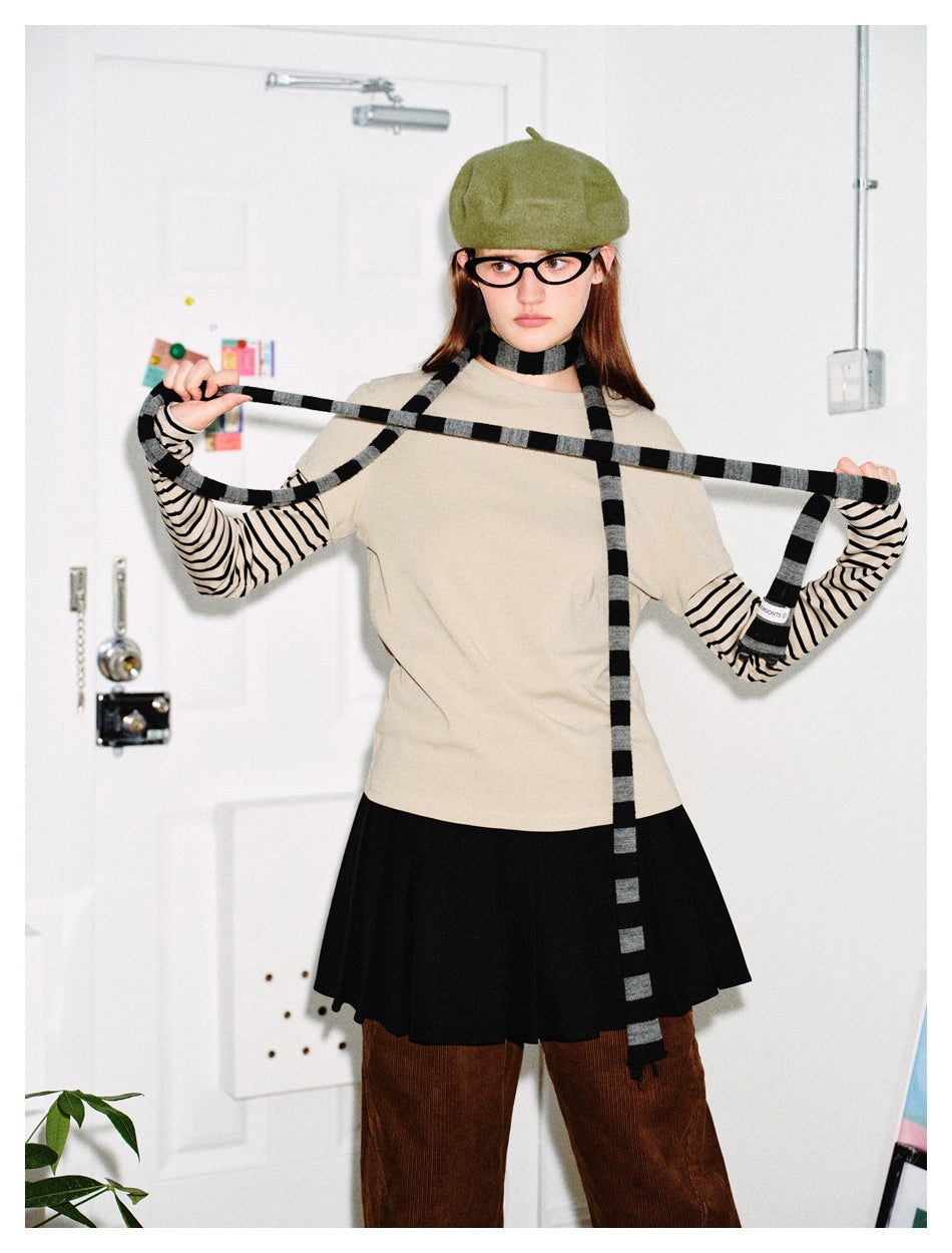 Model holds out a black-and-gray striped scarf over a beige top, striped sleeves and black skirt.