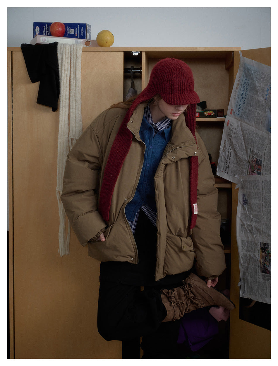 Model wearing a roomy taupe padded jacket with snap details, layered over blue, with a burgundy knit hat and scarf.