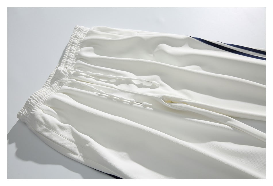 Close-up of white track pants laid flat, showing the elastic waistband, drawstring, and dark side stripe.