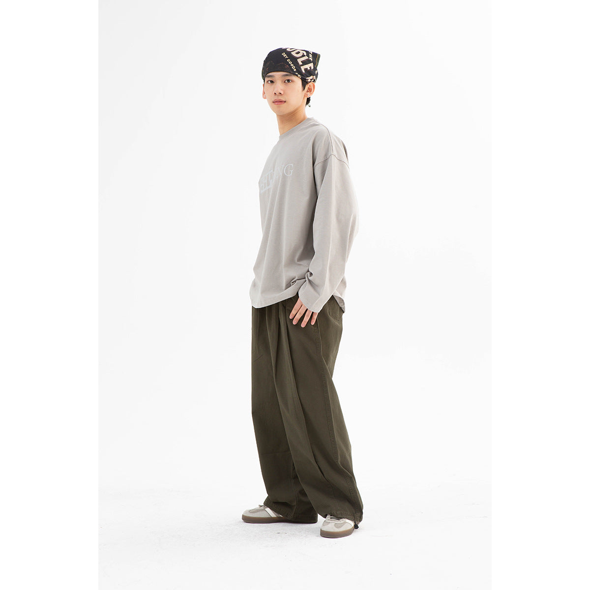 Model wearing a light gray long-sleeve graphic shirt, loose dark olive trousers, and a black printed headscarf.