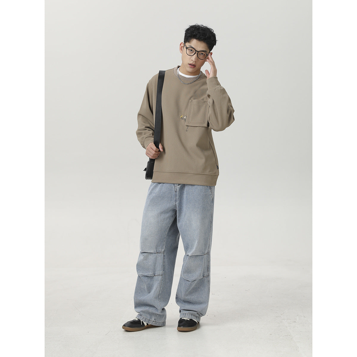 Man posing in a taupe pocket sweatshirt and loose light-wash jeans, with a black crossbody bag.