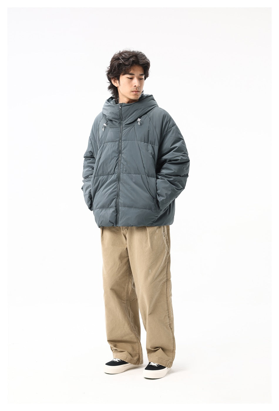 Full-length model wearing a slate-gray hooded puffer jacket and tan trousers, facing slightly to the side.