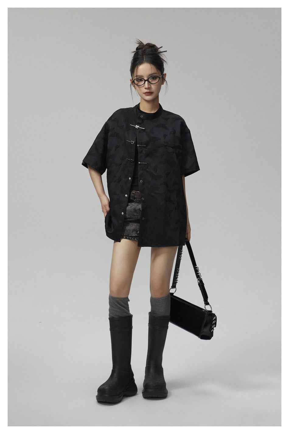 Model wearing the black short-sleeve stand-collar shirt open over a dark top with denim shorts, tall boots and a black bag