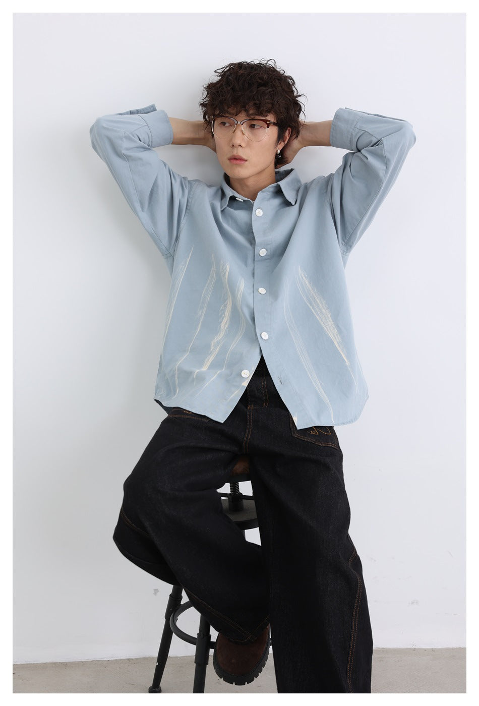 Model wearing an oversized pale blue-gray button-up shirt with white brushstroke graphics and dark wide-leg jeans.