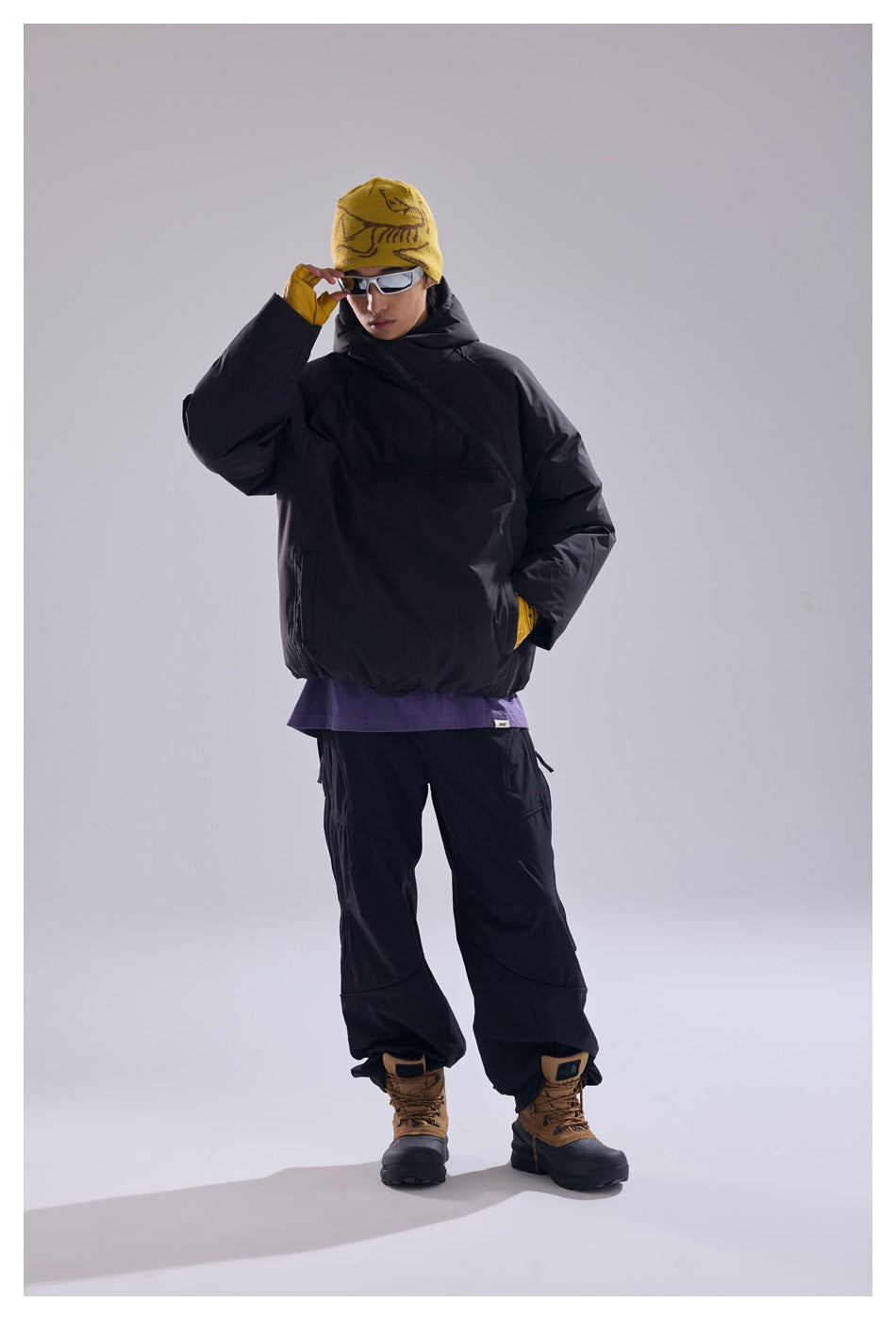 Model wearing a black hooded pullover and black trousers, with a mustard patterned beanie and yellow gloves.