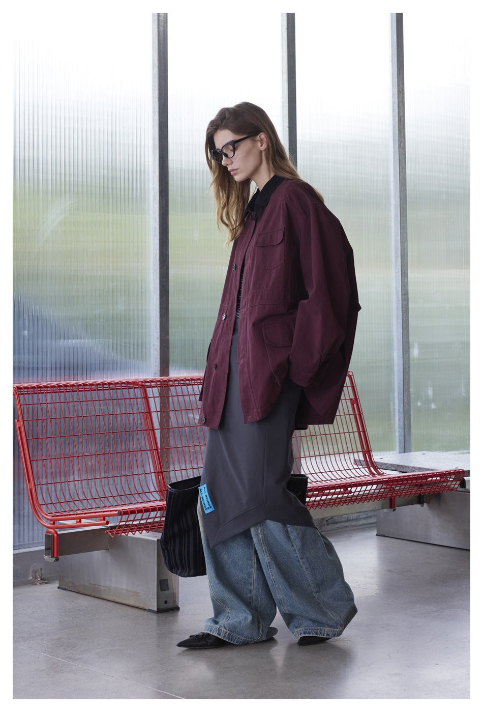 Model in profile wearing an oversized burgundy jacket with black collar, dark skirt, and loose blue jeans.