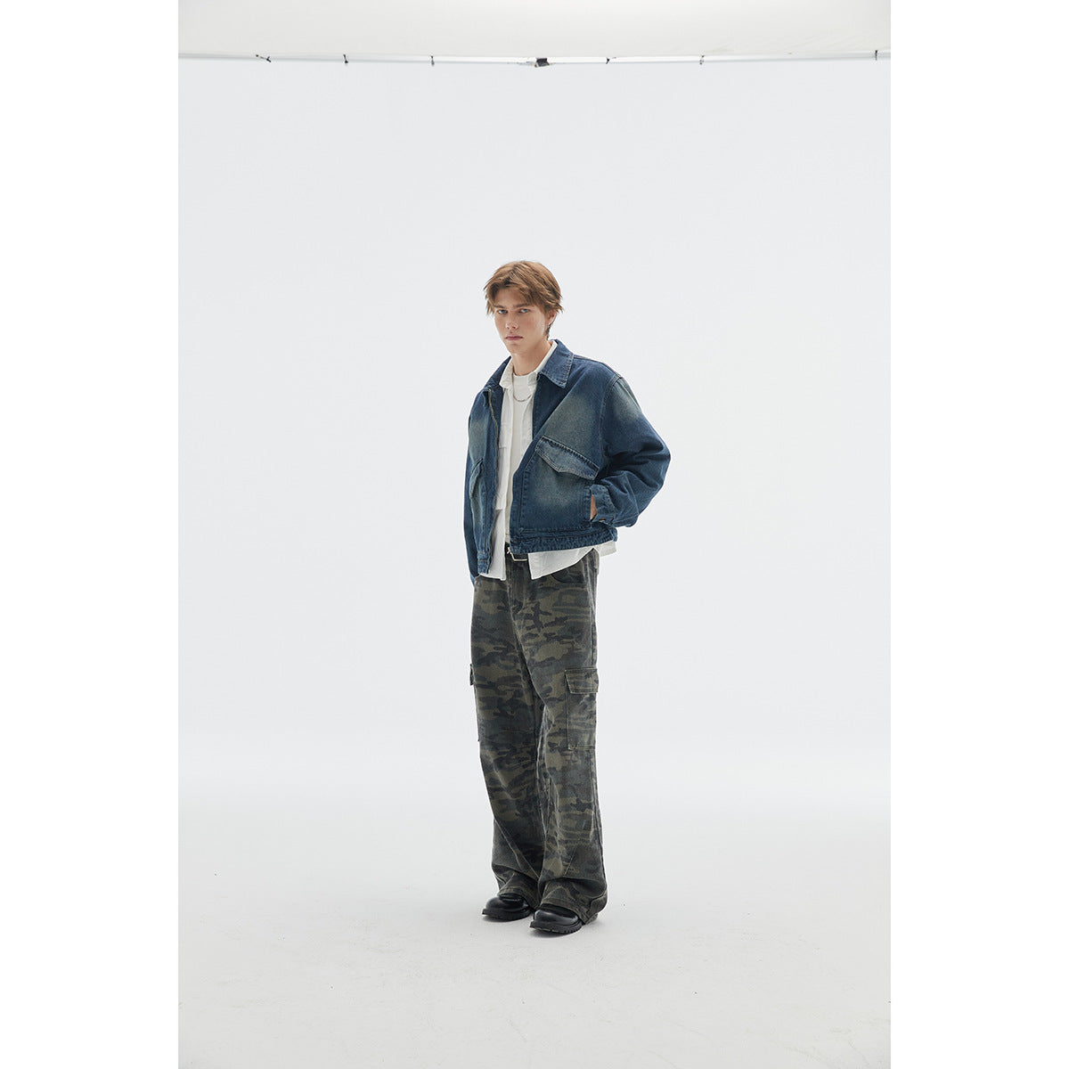 Model in a dark blue denim jacket, white layers, and loose camouflage cargo pants, standing at a slight angle.