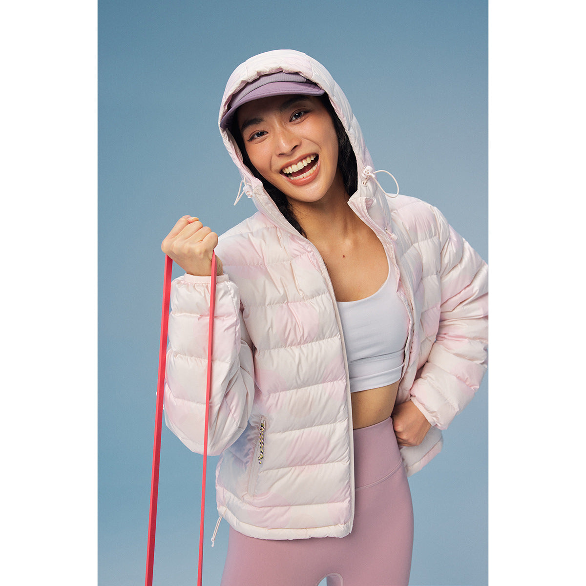 Smiling model wearing an open pale pink hooded puffer jacket with subtle circular pattern, white top, and lavender leggings.