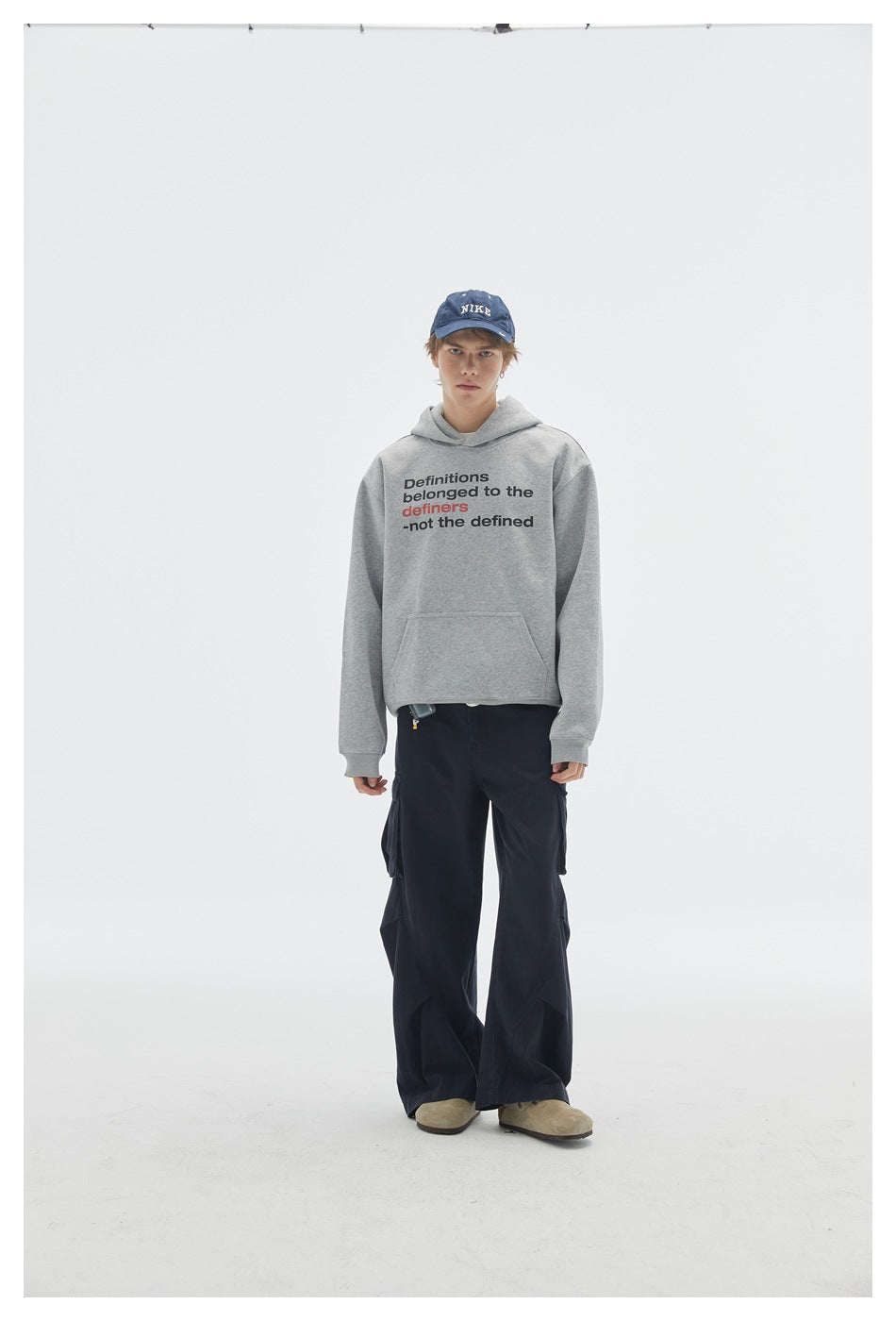 Model wears a gray slogan hoodie with kangaroo pocket, navy cargo pants, and blue cap, shown full-length facing forward.