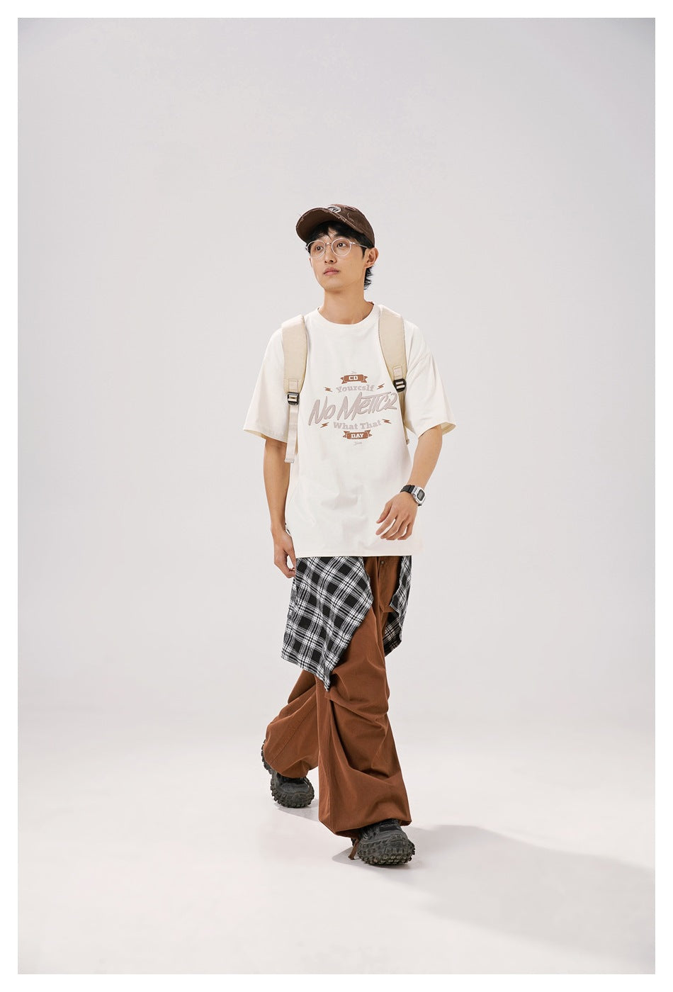 Model wearing an ivory graphic T-shirt with a beige backpack, brown trousers and black-and-white plaid fabric.