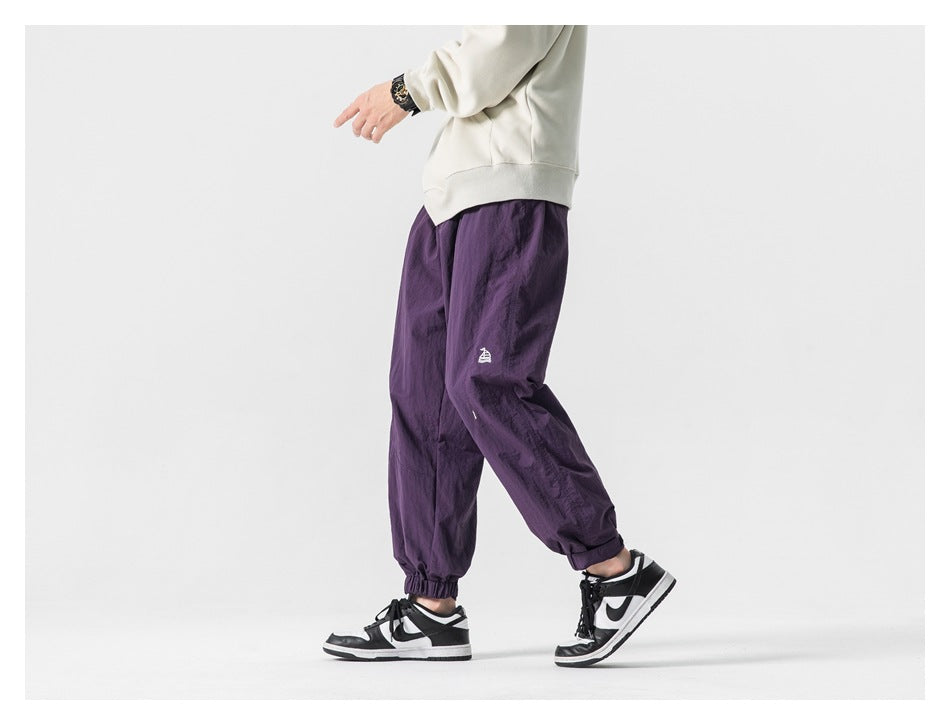 Side view of a model wearing deep purple loose-fit jogger pants with elastic cuffs and a small embroidered emblem.