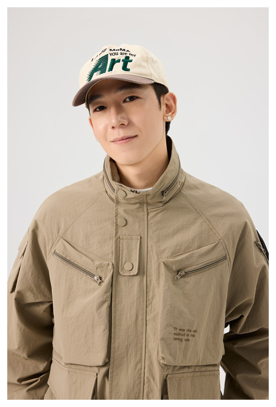 Close view of a man wearing a tan utility jacket, showing its high collar, snap placket, zip chest pockets, and sleeve patch.