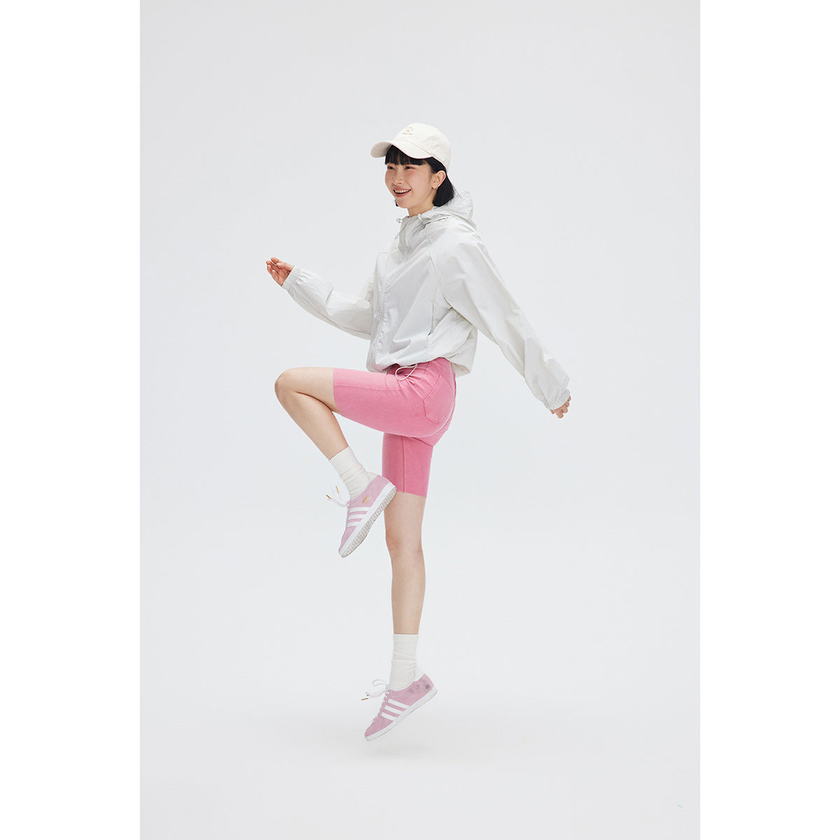 Model in a white hooded jacket and pink cycling shorts, posing mid-step with a white cap and pink sneakers.