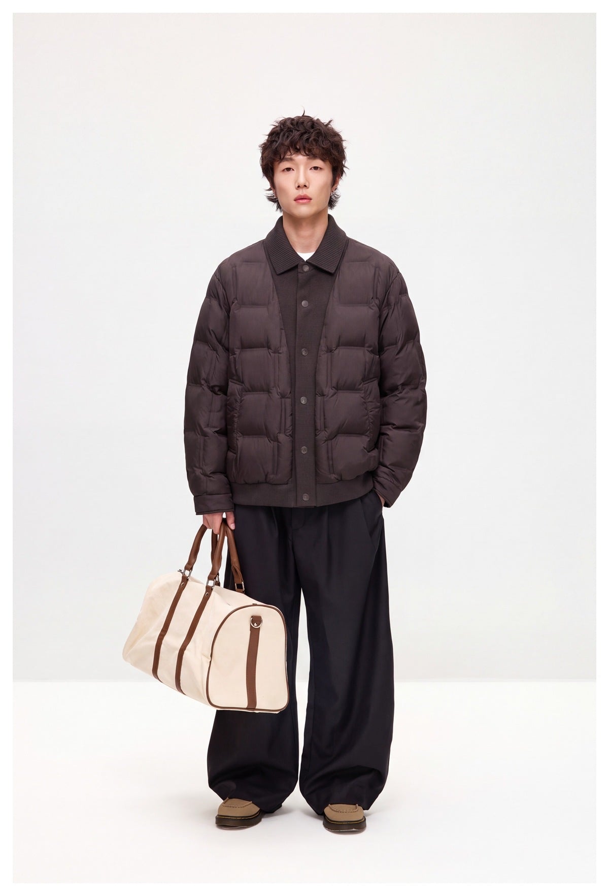 Model wearing brown quilted jacket with black wide-leg pants, holding a cream and brown duffel bag, full-length view
