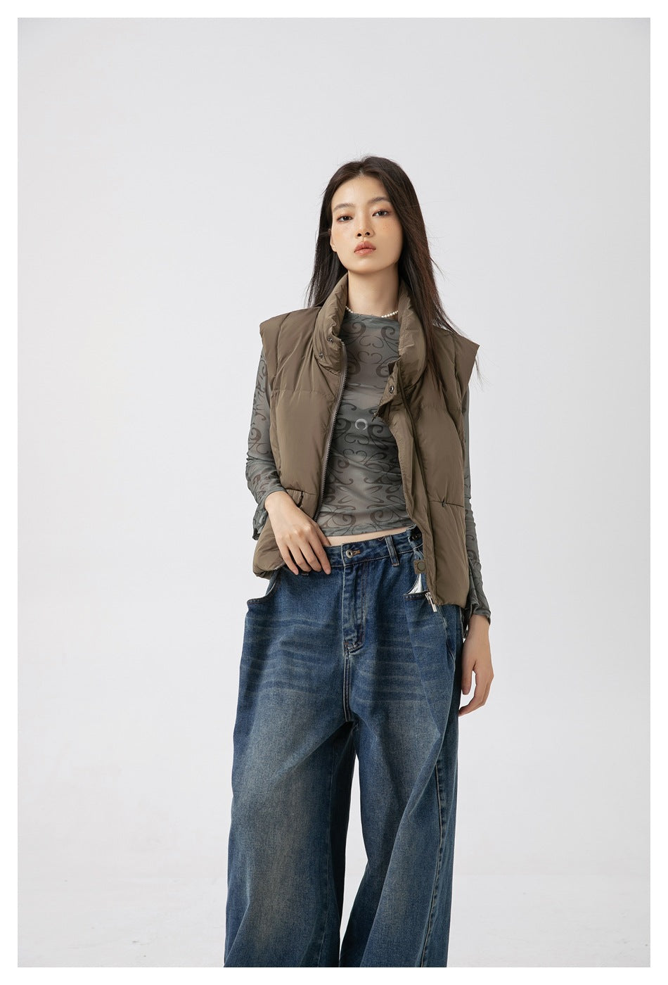 Model wearing an open taupe padded vest over a gray patterned sheer top and wide-leg blue jeans.
