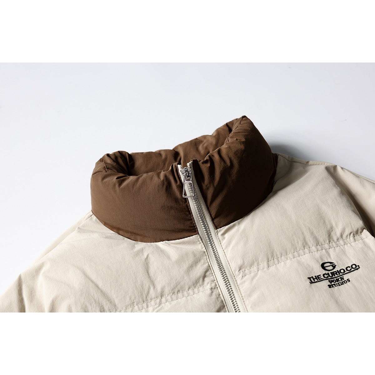 Close-up of a cream jacket with a padded brown collar, silver zipper and black chest embroidery.